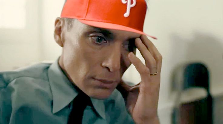 A scene from the movie Oppenheimer shows Cillian Murphy as J. Robert Oppenheimer, sitting in uniform with a pained expression, hand on his face. A red Philadelphia Phillies baseball cap has been digitally added to his head, creating a humorous contrast.