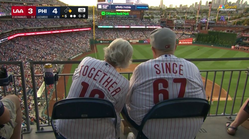 An elderly couple sits together at a Phillies baseball game at Citizens Bank Park, overlooking the field from the outfield stands. They are wearing matching Phillies jerseys: the woman’s reads “TOGETHER 19” and the man’s reads “SINCE 67,” symbolizing they've been together since 1967. The stadium is packed with fans, and the Philadelphia skyline is visible in the background during sunset.