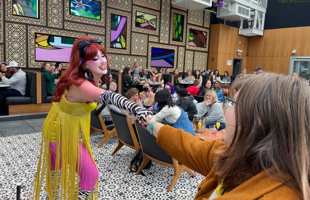 Gorgeous drag queen Kylie in a yellow-fringe dress, zebra gloves and boots and hot pink tights taking a tip from my gorgeous daughter at a drag brunch