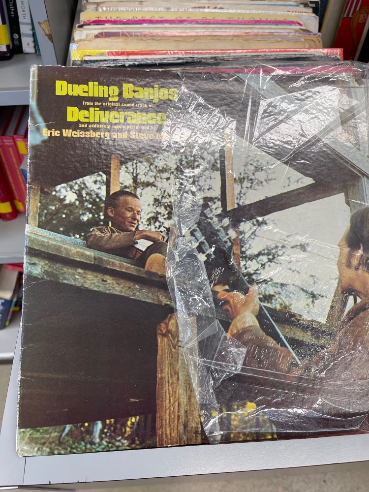 A record album called Dueling Banjos from the movie Deliverance showing one banjo player sitting in a raised wooden structure, slightly elevated while another banjo player faces him below.  Creepy af.