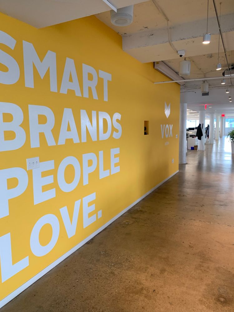 An empty office. A large wall in the foreground is painted in yellow, with white letters overlaying, with the Vox Media logo and the slogan SMART BRANDS PEOPLE LOVE.