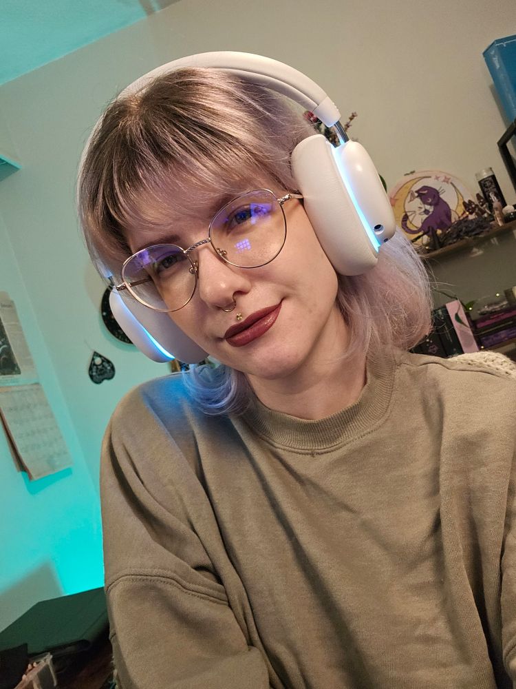 A selfie of Ash with her silver, shoulder length hair down. They are looking at the camera with a soft gaze and slightly smiling. She's also wearing a cozy looking green crew neck sweater.