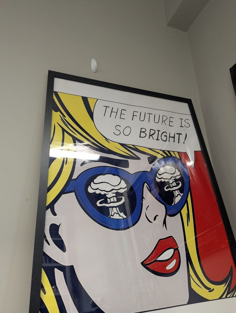 A Roy Lichtenstein print showing a woman in sunglasses painted in a cartoon style. In her sunglasses is the reflection of a nuclear bomb cloud. 
The print is in a black frame leaning against a grey wall with a white hook above. 