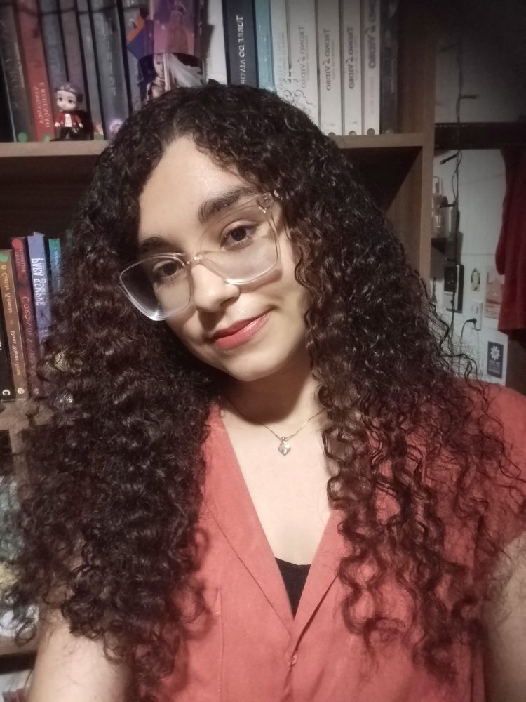 De fundo uma estante, de frente uma selfie minha. Pele branca bronzeada, óculos quadrado de armação transparente, cabelo cacheado longo e castanho escuro, olhos castanhos escuros, camisa laranja de botões 