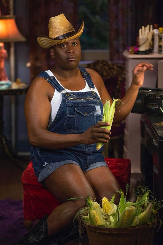 Tituss Burgess in the Netflix show Kimmy Schmidt. He’s shucking corn while wearing denim overalls—cut into short shorts with a white tank top underneath and in a cowboy hat. 