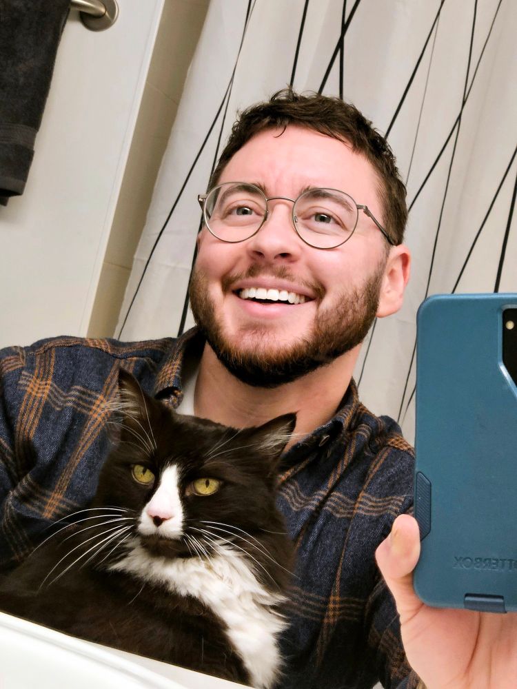 Corin (a white man wearing glasses and a plaid shirt) taking a bathroom mirror selfie and laughing with a gorgeous dark brown and white longhaired tuxedo cat in his lap
