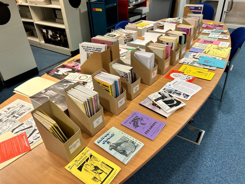 A full table of archive material and special collections publications showing small run community publications and erotic zines from predominantly the 1970s, as well as catalogues in the background and 1980s-1990s club flyers.