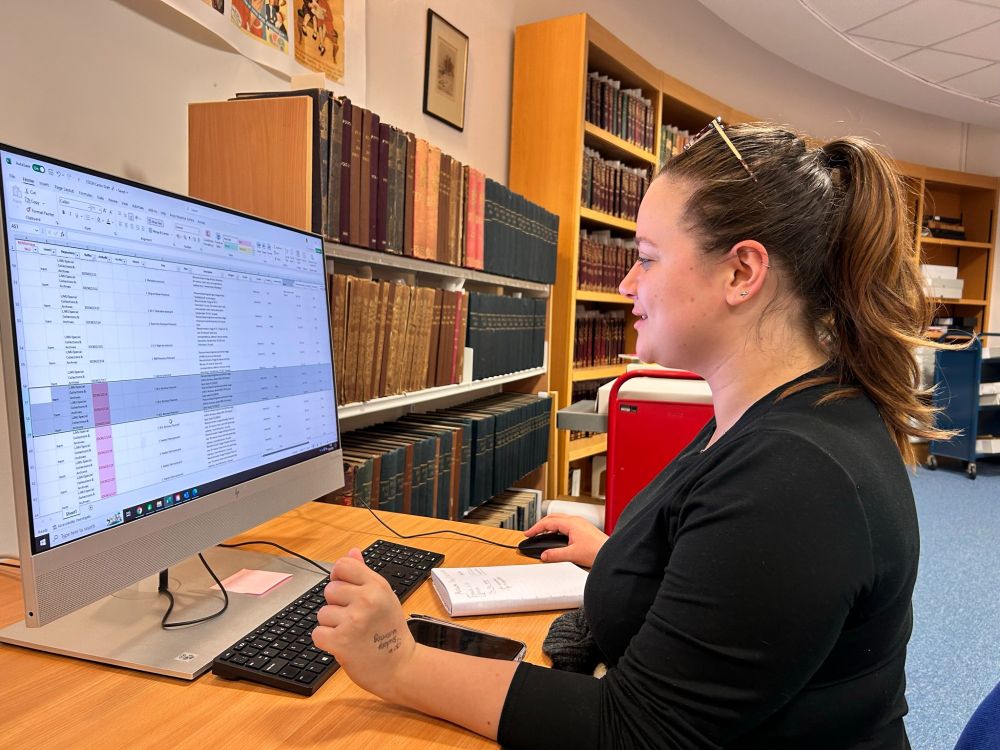 A white female student sits at a desk looking at a screen with an Excel catalogue up. She is wearing glasses on her top of her head, and has a brown ponytail. Behind her is our Reading Room, and shelves of 19th Century periodicals.