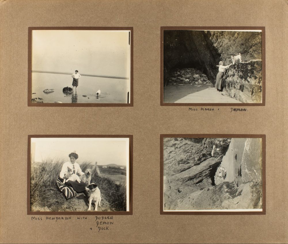 Four photographs pasted into a photograph album page. The first shows Henderson in the sea, hoisting up her skirt, with one terrier dog. The second shows Marsh on a sandy bank near some large rocks, gesturing to Demon the terrier dog who has climbed up onto a ledge. The third shows Henderson sat down in the dunes with a union jack pillow, with all three terrier dogs Dodger, Demon, and Dock. The fourth is a photo of some cliff ledges on the sandy bank, taken from above.