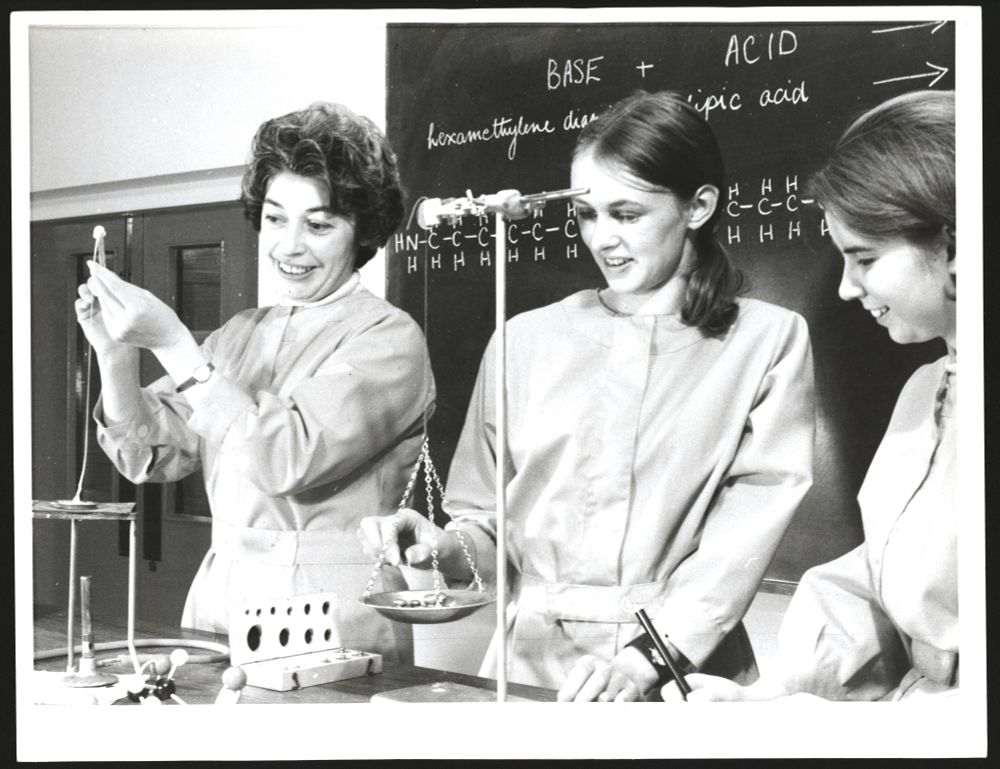 Black and white photo of three white women in lab coats investigating the properties of nylon. One is weighing something on a balanced weighing scale, while another is pulling up a thread of melted presumed-nylon. Behind them is a blackboard with chemical formulas written in chalk.