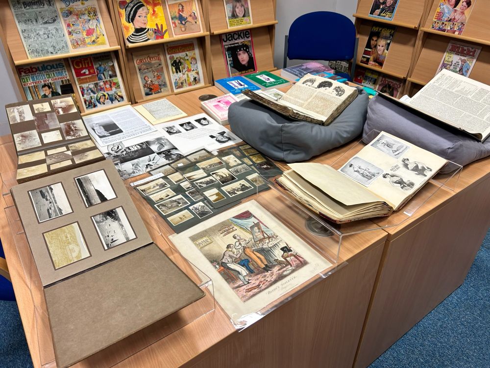 A table full of archive material and special collections publications, including photograph albums from I M Marsh College, a satirical book cutting mocking dandies, two scrapbooks of 19th century period women's costume, The Family Doctor, some of Peter Farrer's publications, & etc.