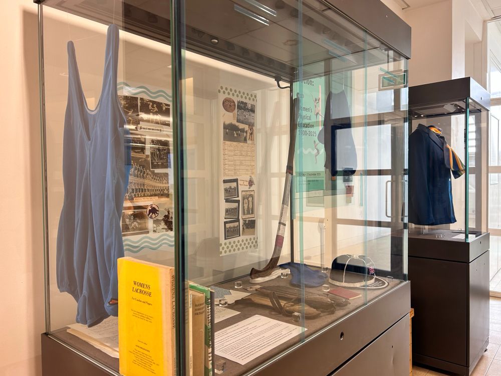 A photo of two exhibition cases. The closest has blue transparent vinyls down the front to split the case into three 'lockers.' On one side is a hanging swimming costume, some books, and some interpretation panels with photos and text, and on the other side is a hockey stick, a hockey cap, some shin pads, a small red book, and some hanging socks with a field sports interpretation panel at the back. In the background you can also see another tall exhibition case featuring a headless mannequin wearing an I M Marsh uniform scarf and blazer. In between the two cases is a bench to sit on, and the header poster for the exhibit.