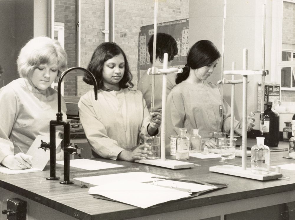 Black and white Photo of Students Doing Science, Testing the Hardness of Water. Three students, one white, one possibly South Asian, and one possibly East Asian, stand at a lab bench with beakers and other lab equipment, making notes.