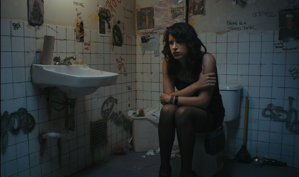 Shirin looking upset and sitting on the toilet. The bathroom walls are covered in graffiti. 