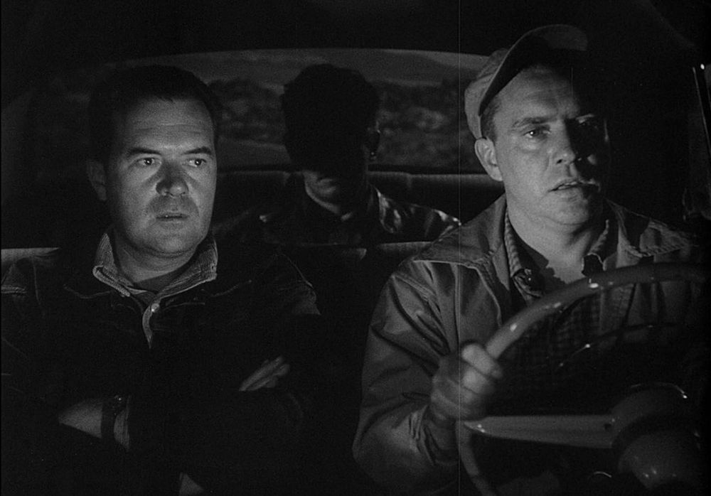 Black and white photo of two middle-aged men looking alarmed and sitting in the front seat of a car. Another man is sitting in the backseat, his face obscured by shadows. 