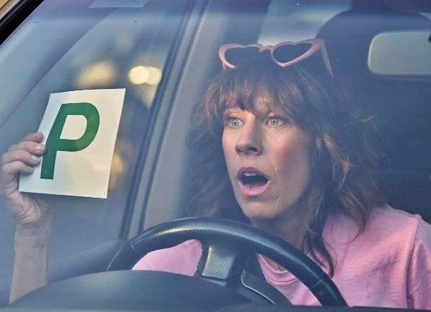 Ronnie pretending to be Audrey wearing heart-shaped sunglasses on her head and holding up a "P" plate indicating that she is a new driver.