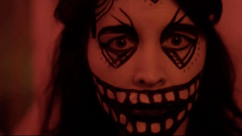 Alice Lowe as Ruth in Prevenge wearing white makeup with black details over her mouth and eyes to appear like a stylized skull. The lighting gives a blood red effect to the frame.