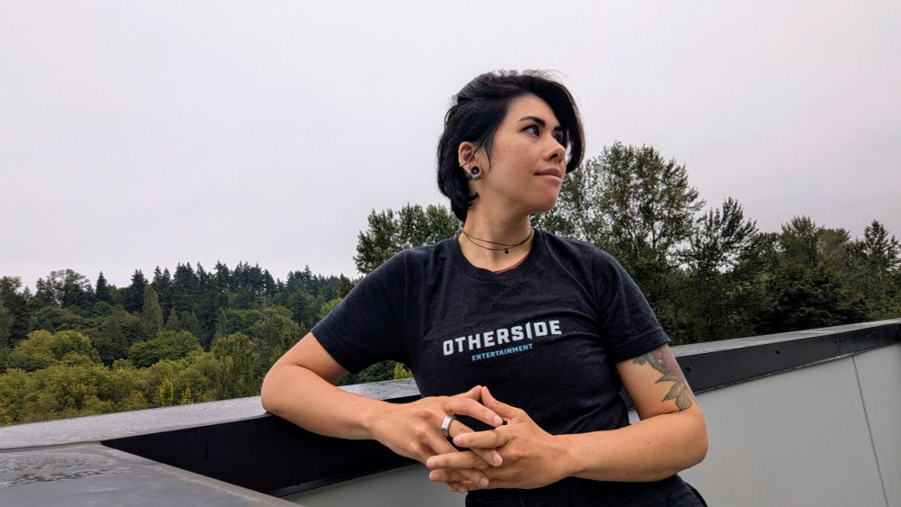Ellie leans against an industrial railing, smiling at something in the middle distance. They wear a brand-new shirt with the words "OTHERSIDE ENTERTAINMENT" emblazoned across the chest. Their hands are lightly laced in front of them, ready to get to work.