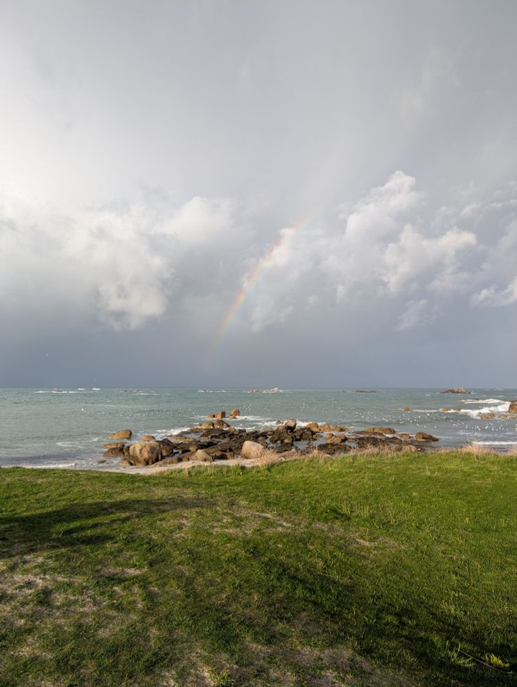 Une vue de la mer, avec une pelouse verte au premier plan, des rochers au milieu et un arc en ciel en plein milieu, devant un ciel très chargé. La mer est gris-bleu.