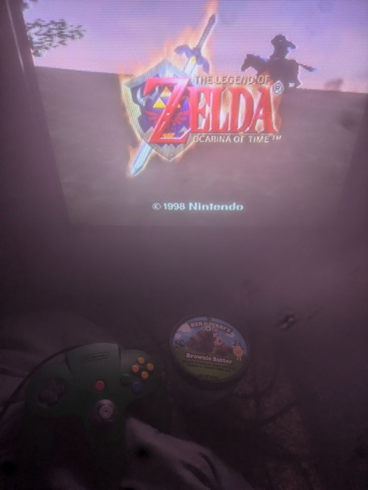 an image of a crt displaying the zelda ocarina of time title screen. there is a pint of ben and jerrys brownie batter ice cream on the floor next to a green nintendo 64 controller. The crt is named tom, and the controller is named bernard.