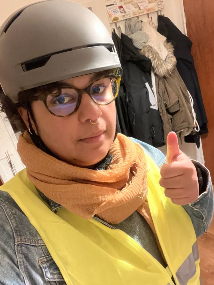 Shelly trägt eine blaue Jeansjacke, darüber eine gelbe Sicherheitsweste, einen gelben Schal, eine braune Brille und einen grauen Helm. Hen schaut recht neutral in die Kamera und zeigt den linken Daumen hoch 👍🏽 