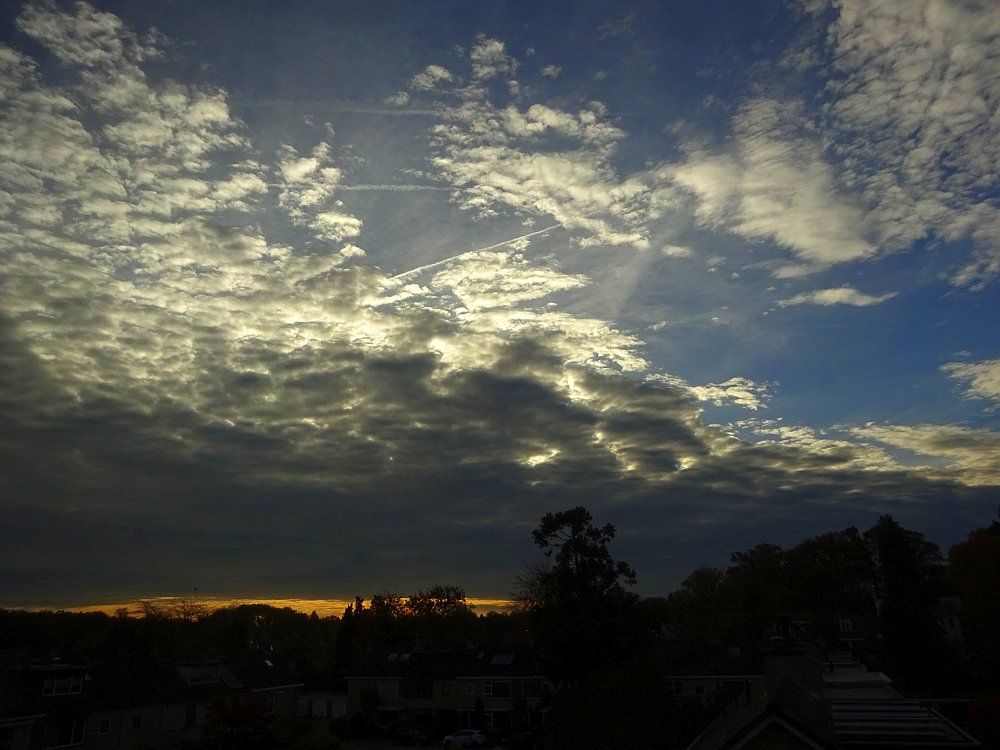 Weer thuis een blik uit het keukenraam. De woonwijk is nog donker, er is een gele streep aan de horizon, daarboven donkere wolken waarvan de bovenkant fel oplicht, rechtsboven blauw lucht. 

Back home, a look out the kitchen window. The residential area is still dark, there is a yellow line on the horizon, above it dark clouds with the top glowing brightly, blue sky at the top right.