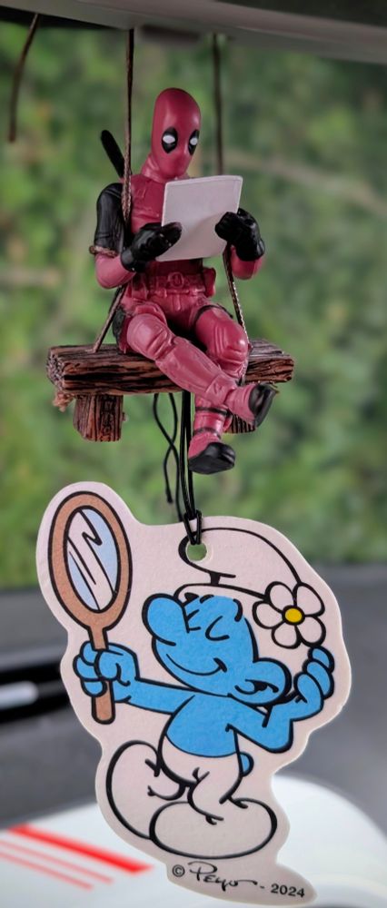 Das Bild zeigt einen Deadpool-Figurenanhänger auf einer Schaukel, der ein Blatt Papier liest, sowie einen Schlumpf-Figurenanhänger, hier Beauty Schlumpf, der einen Spiegel hält.

Das Bild zeigt zwei Figuren-Anhänger, die an der Innenseite dea Autorückapiegels befestigt sind. Oben hängt ein Deadpool-Figurenanhänger, der in einem roten und schwarzen Anzug auf einer kleinen Holzbank sitzt, die an einer Schnur befestigt ist. Deadpool hält ein weißes Blatt Papier in seinen Händen und scheint zu lesen. Der Ausdruck in seinen Augen ist ruhig und konzentriert.  Die Holzbank wirkt einfach und rustikal. Der Hintergrund ist unscharf, zeigt aber eine grüne, natürliche Umgebung.

Unten hängt ein Schlumpf-Figurenanhänger, genauer Beauty Schlumpf. Er ist hellblau mit weißer Kleidung und hält einen ovalen Spiegel in der Hand. Er hat einen freundlichen und sanften Ausdruck, während er selbst verliebt in den Handspiegel blickt.  Die Farben des Schlumpf-Anhängers sind hell und freundlich. Und augenscheinlich ein Duft-Anhänger.

Die gesamte Stimmung des Bildes ist ruhig, etwas humorvoll aufgrund der Kombination von Deadpool und einem Schlumpf, und gleichzeitig liebenswert. Der Kontrast des eher düsteren Deadpool mit dem freundlichen Schlumpf erzeugt eine interessante Dynamik. Das Bild wirkt wie ein Schnappschuss einer persönlichen Dekoration in einem Auto.
