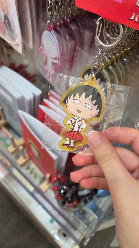 Photo of a keychain, showing a nervous looking school girl character. The character is wearing a white shirt and a red skirt.