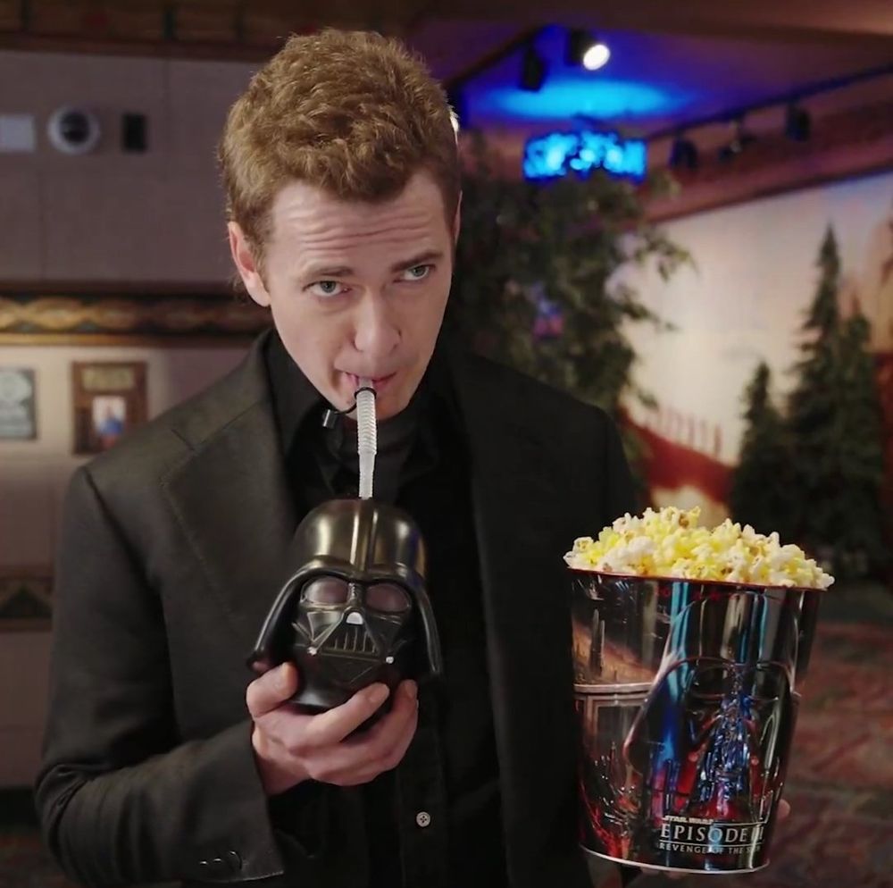 Screenshot of actor Hayden Christensen (who plays Anakin Skywalker/Darth Vader in Star Wars) holding a "Revenge of the Sith" popcorn bucket. He is leaning down to sip on his Darth Vader Helmet Cup. He has a bit of a smug look on his face. He is standing in a cinema, the background is blurred though. You can make up a carpet some plants and a ceiling with some lights. 