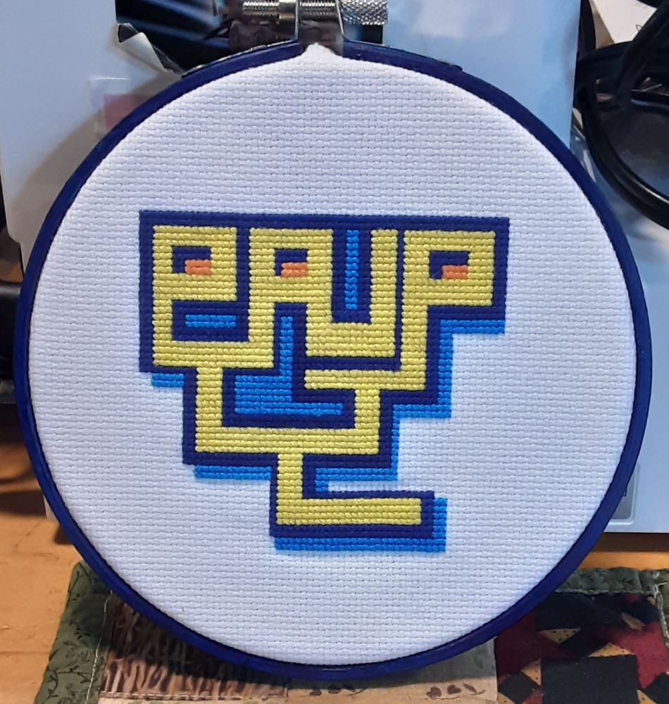 A cross-stitched logo for the phylogenetic inference program PAUP. In yellow are the letters P-A-U-P, attached at the base in the pattern of a phylogenetic tree. The "tree" is outlines in dark blue, with light blue in the background. The counters in the Ps and A are orange.