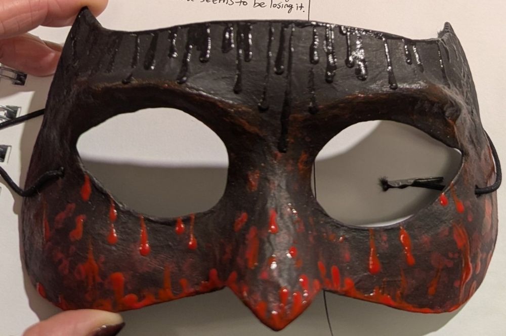 A Halloween half face mask inspired by the masque of the red death by Edgar Allen poe. It is black and drippy with dimensional puff paint on the top, with blood red dimensional paint droplets under the eyes going to the nose. The tops of the corners of the forehead come to points like impish horns.