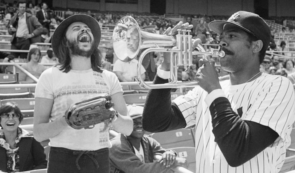 Dock Ellis of the New York Yankees playing around with Chuck Mangione's trumpet while Mangione laughs in the background wearing a baseball glove and a fedora.  Courtesy https://www.semissourian.com/world/jazz-legend-chuck-mangione-known-for-feels-so-good-dies-at-84-a795d6fb