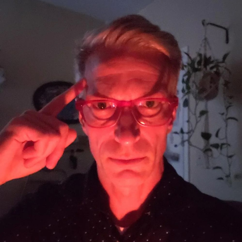 Picture of a man with red glasses on. He Has a finger placed on the side of his head as he stares forward. The lighting is red, making his face look red.
