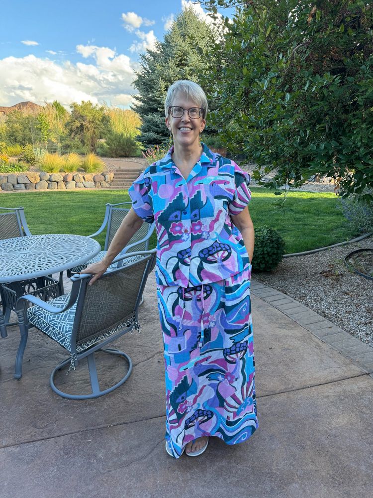 short-haired woman standing outside on a patio wearing a gauzy matching top and skirt in a wild, colorful pattern.