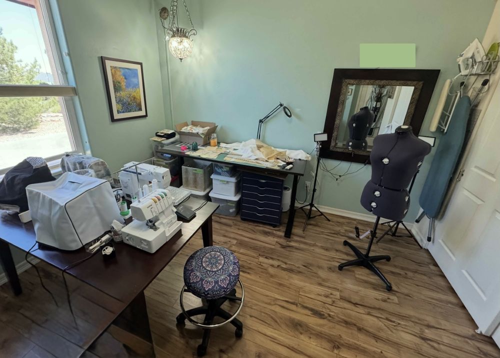 a sewing room with a few machines on one table and cut out fabric on another table.  green walls, brown floor, hanging ironing board and iron.  Lots of lamps.