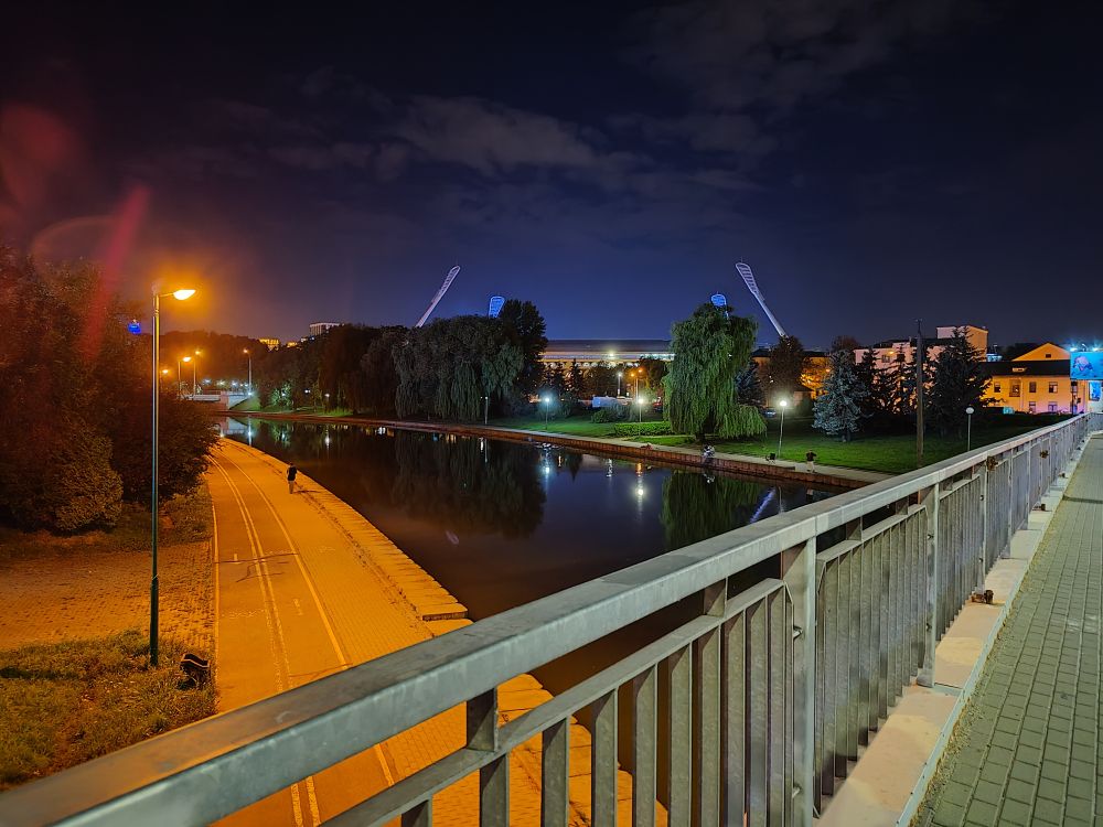 Evening in Minsk, Belarus in August 2025. Bridge over the Svislač river. A lone person is walking on the closer bank, lit with an orange streetlight, and the renovated Dinamo Stadium with swatter-like floodlights is visible on the other bank in the distance. 
