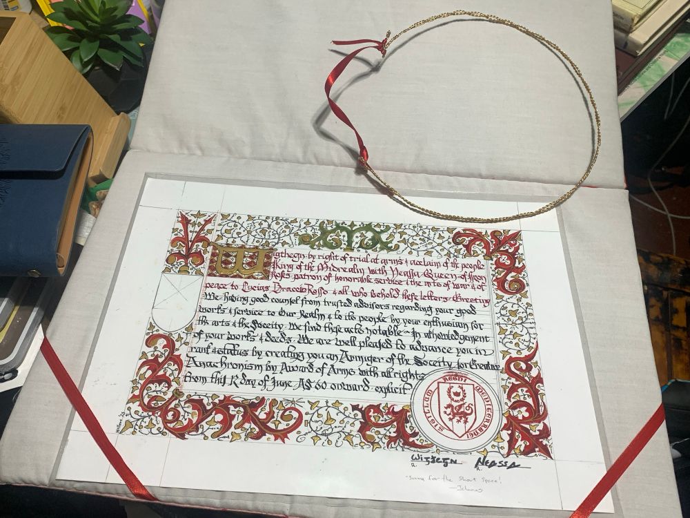 a thin decorative gold wire circlet sized to fit on a head, open at one end with the ends connected by a red ribbon to allow size adjustment; below, a diploma-sized paper is inscribed with medieval-style calligraphy and bordered intricately in primarily red and gold