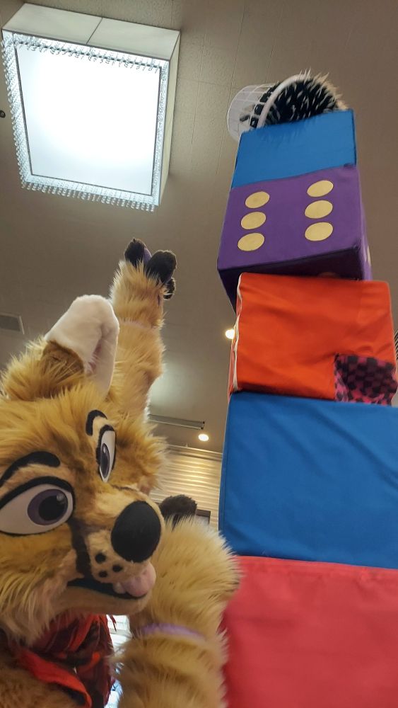 A stack of fabric covered foam blocks rises towards a high ceiling. Atop the blocks rests a fuzzy ball on which a round plastic basket has landed at a precarious angle. Niko points up at the precariously positioned basket from the lower left of the photo in full body fursuit.