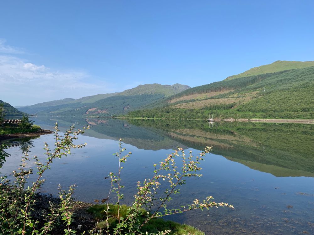 A view across Loch Long. Blue sky. The Arrochar Alps are perfectly reflected in the still water.