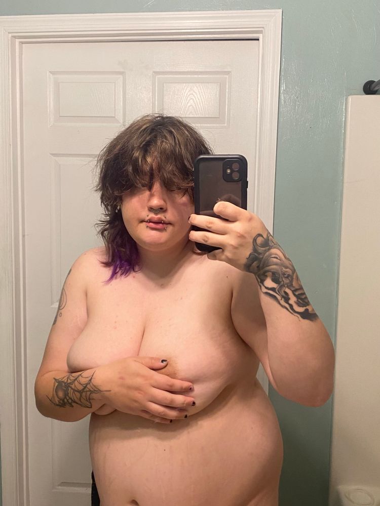 fat genderfluid person posing in a bathroom mirror. they have a shaggy, mid length haircut with purple on the underside. their boobs and belly are visible but are covering their nipples with their right hand and arm. they have multiple lip piercings, arm tattoos, a spiderweb septum ring, and eyebrow piercings.