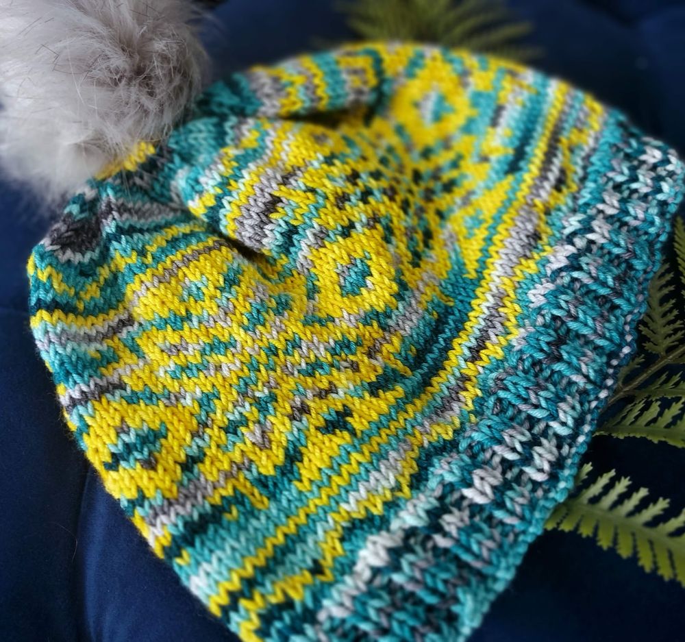 Angled shot of a hand-knit beanie featuring an intricate stranded colorwork pattern in vivid yellow, deep turquoise, and touches of gray and white. The design includes bold geometric and starburst motifs. The beanie has a snug ribbed brim in turquoise and white, and it's topped with a fluffy, light gray faux fur pom-pom. The colors used are "Glimmering Gold Caspar" (a bright golden yellow) and "Blue Theban" (a deep turquoise-blue with specks of gray). The hat is photographed against a rich navy background with fern fronds for contrast.
