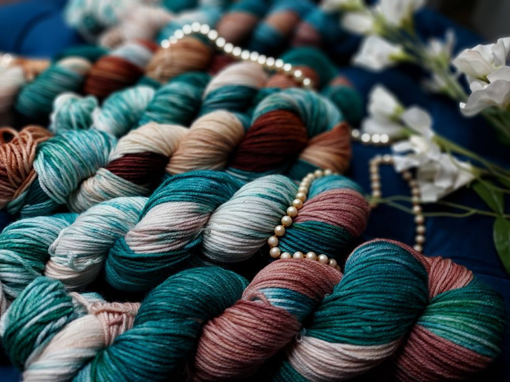 teal, brown, blue and white yarn skein with pearls scattered around and white flowers in the upper right.