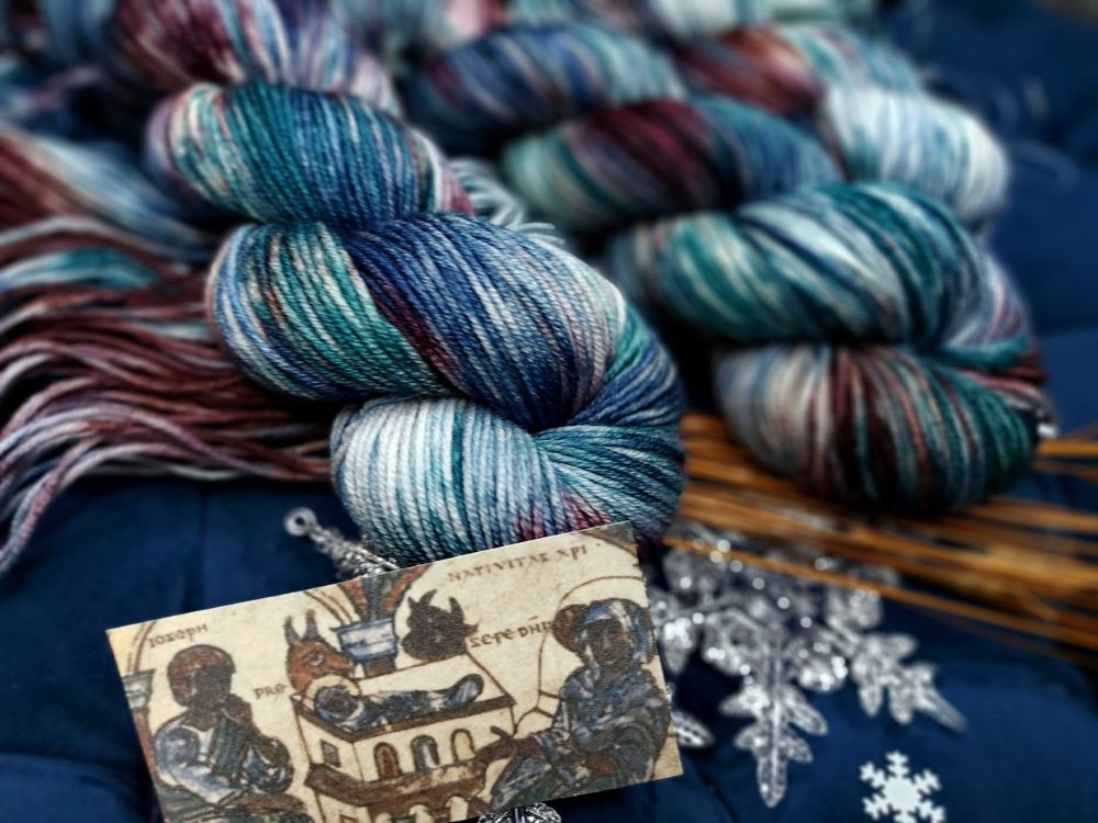 hand-dyed skeins of yarn in a variegated colorway called Midnight in a Manger. The yarn features swirling shades of deep teal, navy, soft cream, rusty red-brown, and denim blue. The skeins are arranged vertically against a dark background with a few small white snowflake decorations. there is an image below the skeins of a medieval nativity scene from a manuscript, depicting the infant Jesus in a rectangular crib, wrapped in blue-striped swaddling cloth. Two people of color on the left and right flank the manger. An ox and donkey peer over the crib, and the illustration is rendered in the stylized, line-heavy medieval style with muted earth-tone pigments.” Holkham Bible Picture Book, a 14th-century English manuscript (sometimes catalogued as Add MS 47682).