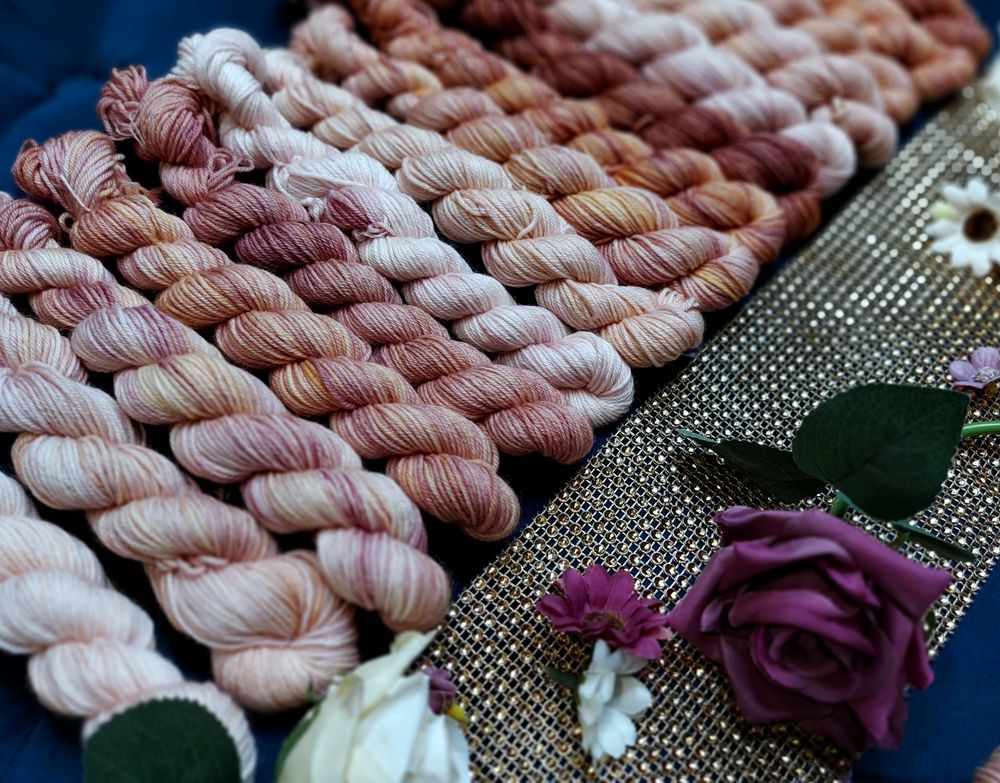 Variegated miniskeins of a rusty plum color, lined up along gold cloth and adorned with purple and white flowers. 