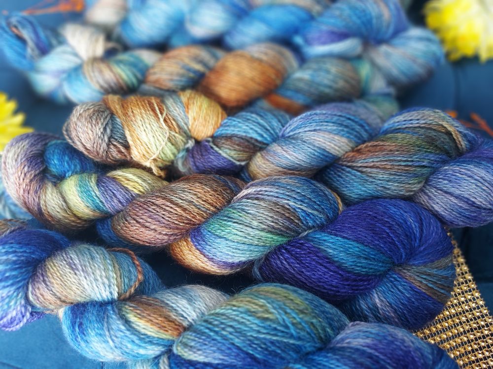 Blue, orange, indigo, soft yellow skeins with faint green in a row. There are orange and yellow dried flowers scattered 