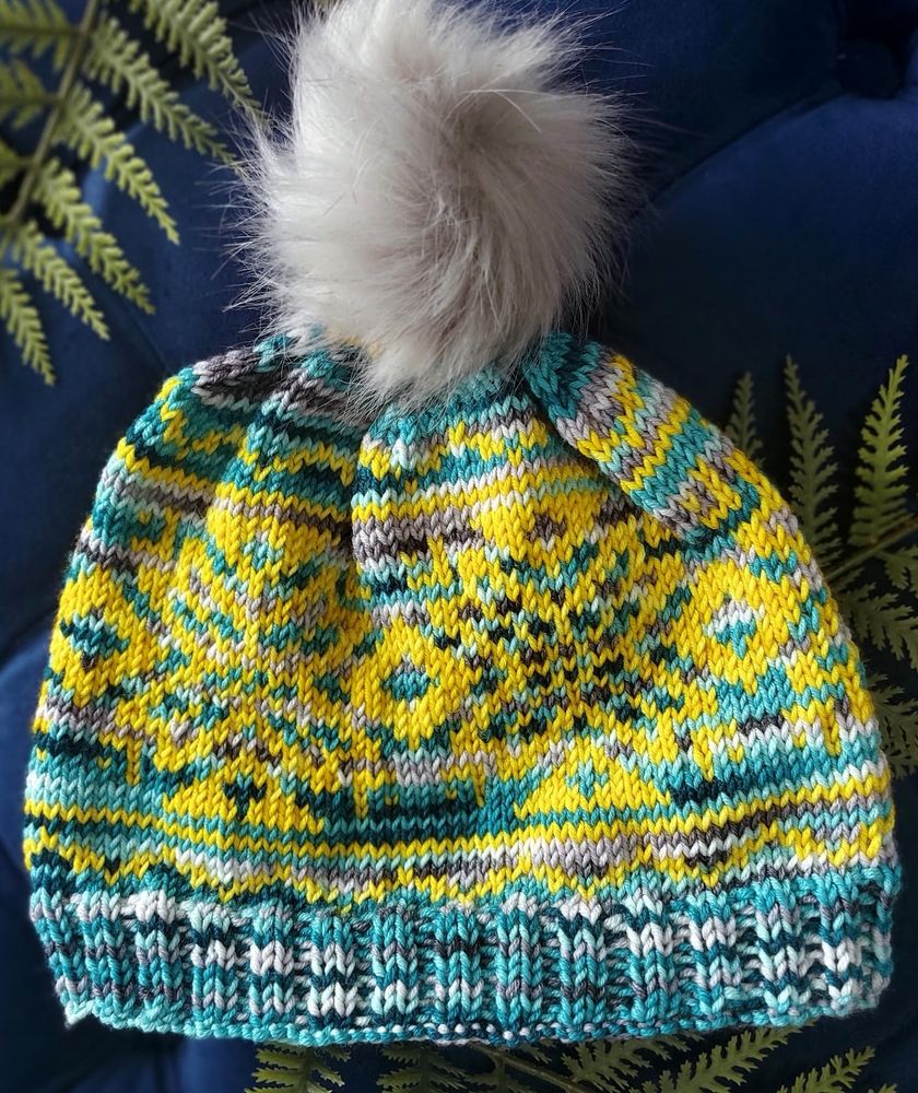 A hand-knit beanie featuring an intricate stranded colorwork pattern in vivid yellow, deep turquoise, and touches of gray and white. The design includes bold geometric and starburst motifs. The beanie has a snug ribbed brim in turquoise and white, and it's topped with a fluffy, light gray faux fur pom-pom. The colors used are "Glimmering Gold Caspar" (a bright golden yellow) and "Blue Theban" (a deep turquoise-blue with specks of gray). The hat is photographed against a rich navy background with fern leaves for contrast.