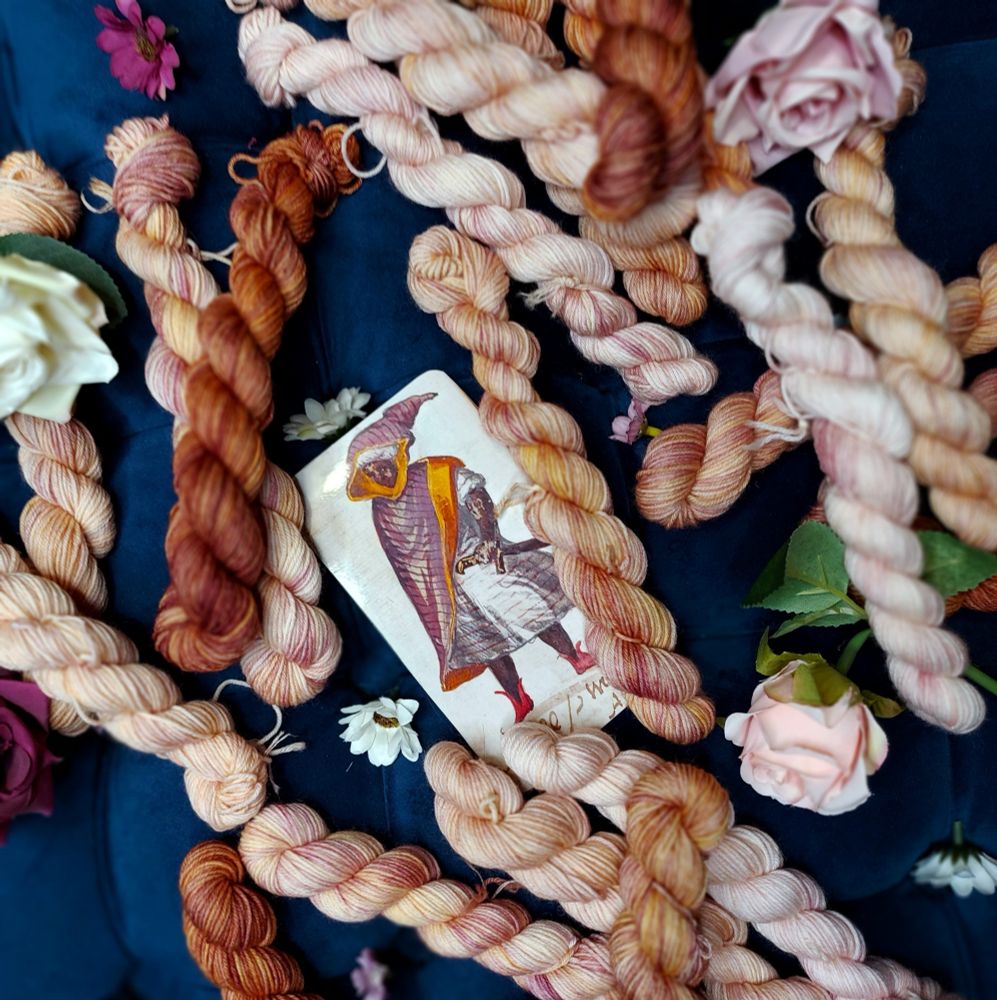 Variegated miniskeins of a rusty plum color, adorned with purple, pink, and white flowers. In the center is a photo of a manuscript depicting a 16th century Black nobleman.