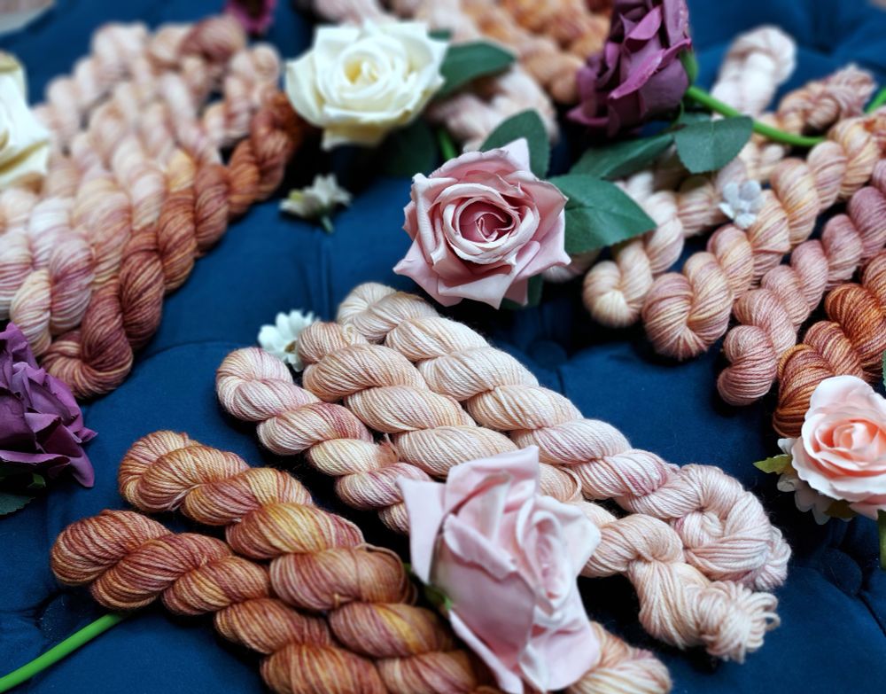 Variegated plum and bronze miniskeins, lined up in bunches and adorned with pink, purple, and white roses.