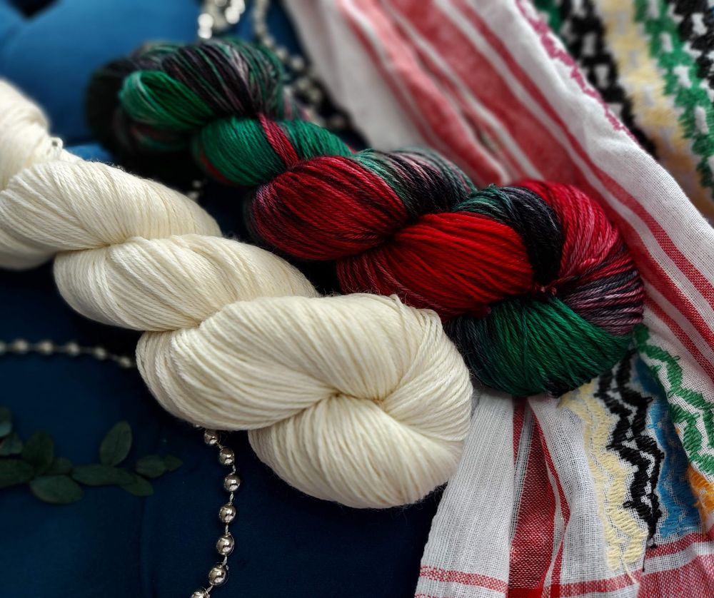 2 skeins of yarn on a rainbow kuffiyeh. There is a creamy white skein and a red, green, and black variegated skein. 