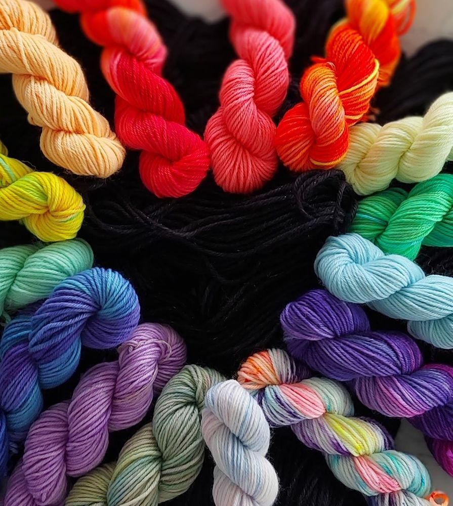 Alt text: A close-up photo of colorful mini skeins of yarn arranged in a rainbow heart on top of black yarn. The skeins include shades of yellow, orange, red, pink, green, turquoise, blue, purple, lavender, sage, and a speckled pastel skein with pink, yellow, and blue. The black yarn in the center creates a striking contrast against the vivid colors.